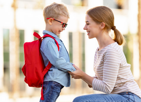 Side view of cheerful woman talking with smart boy with backpack on sunny day outside school...