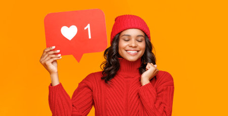 Happy young ethnic female with closed eyes adjusting knitted hat and smiling against yellow...