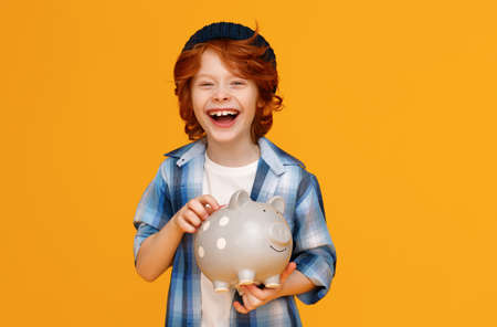 Positive ginger boy in casual outfit laughing and saving money in piggy bank against yellow backgroundの写真素材