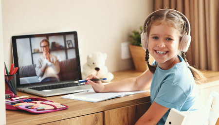 Happy girl in headphones smiling and looking at camera while making video call to teacher during online lesson at homeの写真素材