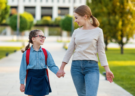 Happy smart girl with notebooks smiling and looking at mother while walking to school in green park togetherの写真素材