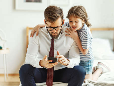 Serious frowned busy man in formal wear and eyeglasses reading business news on smartphone while cheerful little daughter sitting nearby and asking for attention during morning in bedroom at homeの写真素材