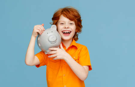 Happy redhead boy in orange shirt cheerfully smiling for camera and carrying piggy bank on shoulder against blue backgroundの写真素材