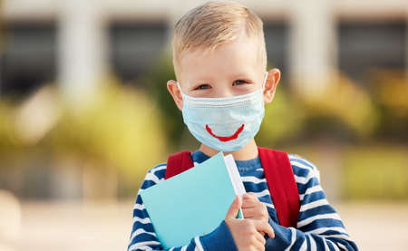 Optimistic smart first grade schoolboy in casual wear and eyeglasses wearing protective mask with drawn smile holding copybooks and looking at camera against blurred urban backgroundの写真素材