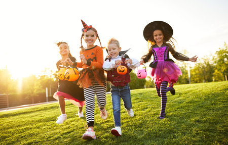 Group of excited kids in spooky costumes smiling and running on lawn during Halloween celebration in evening in parkの写真素材