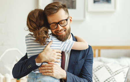 Smiling young bearded man in formal suit and eyeglasses embracing little daughter while sitting in cozy room at home after work dayの写真素材