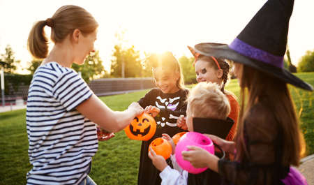 Adult woman giving candies to trick or treating children in costumes during Halloween celebration in evening in parkの写真素材