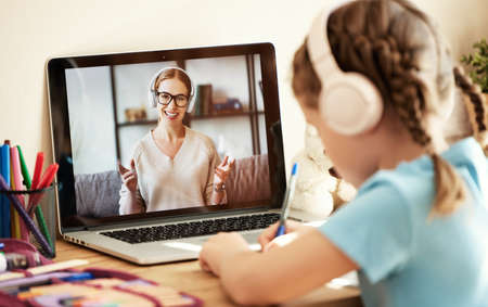 Anonymous girl in headphones sitting at table and listening to friendly teacher smiles and tells interesting stories during online lesson at homeの写真素材