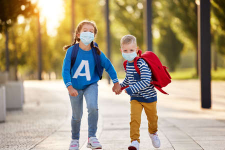 Delighted little sister and brother in casual clothes and protective masks with backpacks holding hands and running together in park ready for school studiesの写真素材