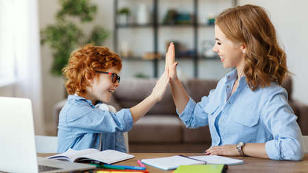 Positive young woman helping son schoolboy in searching information for homework on internet and and giving high five to him while sitting together at table with laptop at homeの写真素材