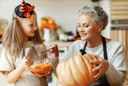 Delighted elderly woman and girl smiling and removing insides of ripe pumpkin while carving jack o lantern for Halloween celebration at homeの写真素材