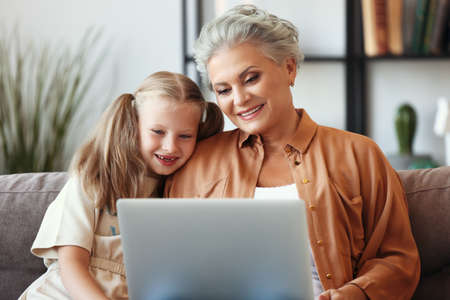 Cheerful senior female enjoying time with little granddaughter while sitting together on sofa and watching funny cartoon on laptopの写真素材