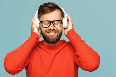 Cheerful bearded man in glasses smiling and listening to good music in headphones with closed eyes...