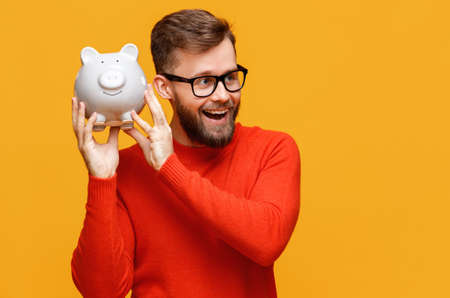 Optimistic bearded man in glasses looking away with opened mouth and shaking piggy bank with money near ear against yellow backgroundの写真素材