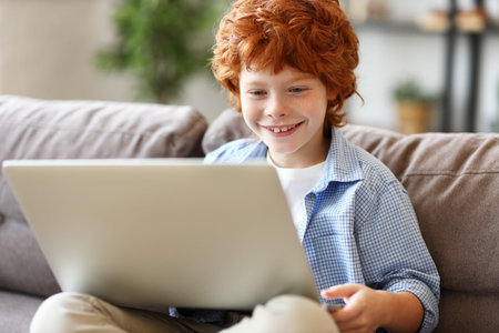 Cheerful redhead boy in casual clothes smiling and playing game on laptop while sitting on soft couch at homeの写真素材