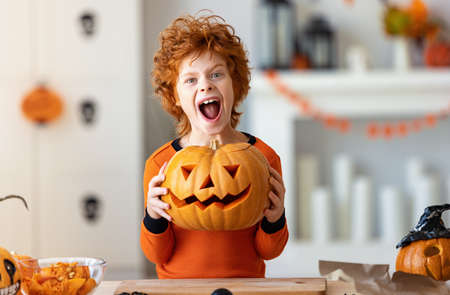 Happy funny ginger boy in a costume with pumpkin Jack o lantern gesticulating while scaring camera during Halloween celebration at homeの写真素材