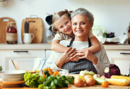 Cute little girl hugging happy grandmother while preparing healthy vegetarian dish with fresh ingredients for lunch in home kitchenの写真素材