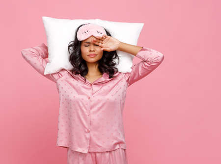 Unhappy ethnic woman in sleepwear and mask holding pillow behind head and rubbing face after early awakening against pink backgroundの写真素材