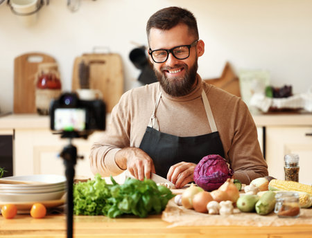 Happy bearded man in glasses smiling and prepare a vegetarian dish ripe vegetables while shooting...