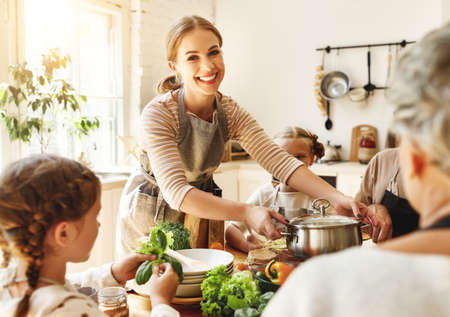 Positive young housewife with little daughters, husband and grandmother gathering around kitchen table and preparing delicious healthy dishes with fresh ingredients for family lunch at homeの写真素材