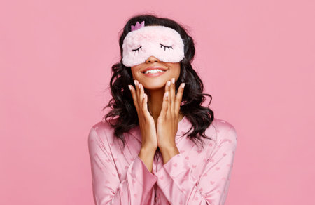 Delighted ethnic female in pajama and mask smiling and smearing lotion on face during skin care routine before sleep against pink backgroundの写真素材