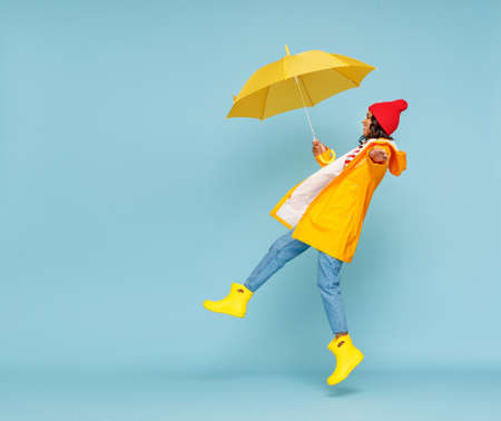 Side view of happy ethnic woman in outerwear and with umbrella leaping and having fun on rainy autumn day on blue backgroundの写真素材