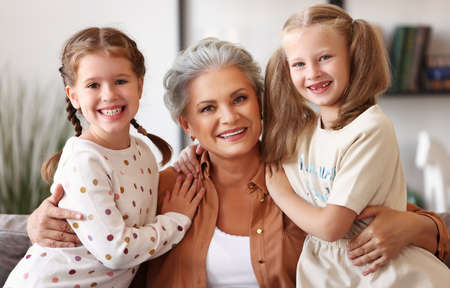 Cheerful senior woman smiling for camera and hugging girls while sitting on couch in cozy living room at homeの写真素材