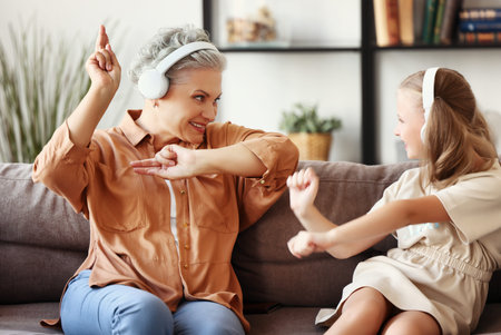 Happy senior woman and girl listening to music in headphones and dancing while sitting on couch at...