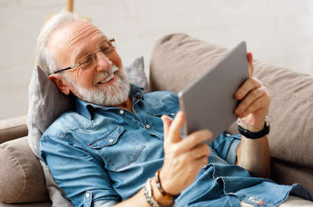 Optimistic bearded elderly male relaxing on sofa and browsing tablet on weekend day at homeの写真素材