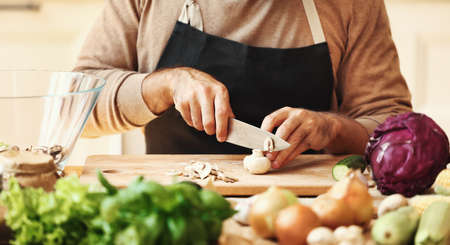 Crop unrecognizable bearded male in apron slicing champignons on wooden cutting board while preparing healthy dish with fresh ingredients in home kitchenの写真素材