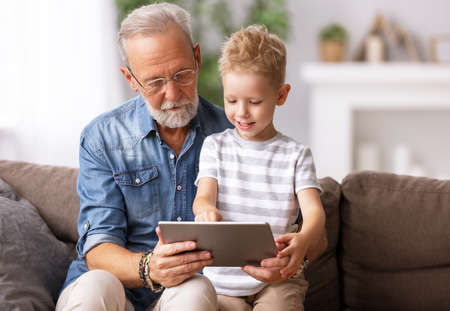 Happy family senior grandfather and boy sitting on sofa and watching cartoon on tablet on weekend day at home togetherの写真素材
