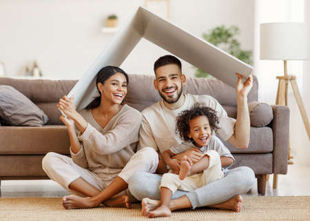 Cheerful parents with child smiling and keeping roof mockup over heads while sitting on floor in cozy living room during relocationの写真素材