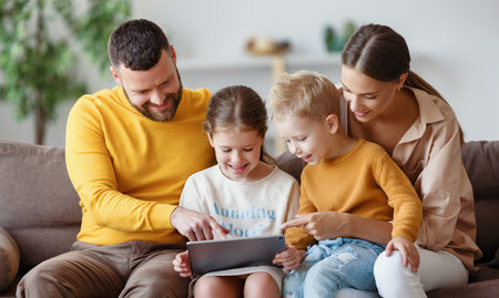 Happy family young parents with smiling children in casual outfit sitting on couch browsing tablet...