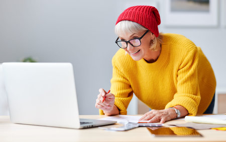 cheerful old woman creative designer in a red hat smiles at workplace at the table with a laptop,...