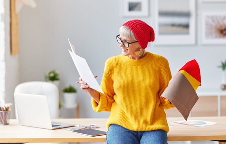 cheerful old woman creative designer in a red hat smiles at workplace at the table with a laptop, sketches and samplesの写真素材