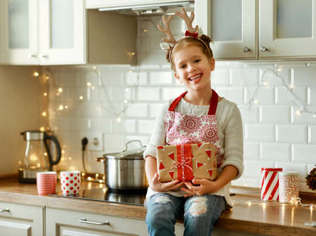 happy funny cute girl with reindeer horns holds a Christmas gift while sitting on cozy kitchen at homeの写真素材