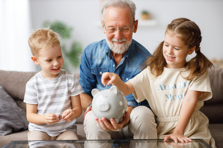 Aged man giving piggy bank to boy and girl while showing grandchildren how to save money at homeの写真素材