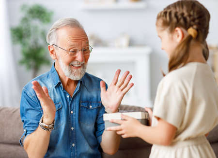 Cheerful elderly male smiling and gesticulating while receiving surprise gift from granddaughter during holiday celebration at homeの写真素材