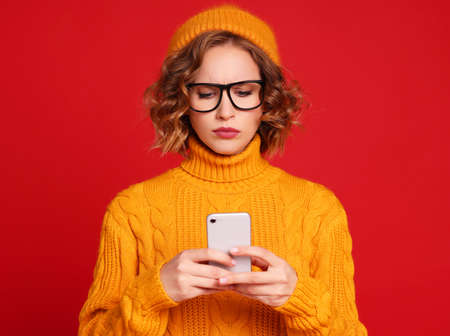 thoughtful young woman blogger in a yellow sweater, glasses and hat exchanges messages with friends using a smartphone on a bright red backgroundの写真素材