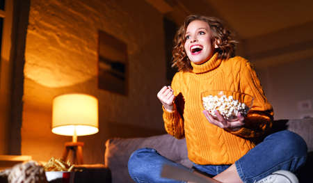young delighted cheerful woman eating popcorn laughs and watches comedy movie on cable TV at home in evening aloneの写真素材