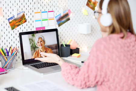 Side view of cheerful young woman in pink sweater smiling and speaking with mother while sitting at table and making video call at homeの写真素材