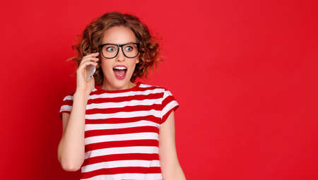 Amazed young female with curly blond hair looking away while talking on mobile phone against red backgroundの写真素材