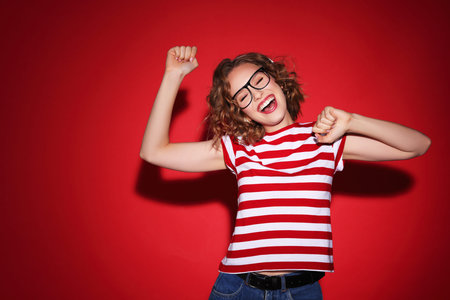 Happy young woman in striped t shirt and stylish glasses smiling and dancing against red background