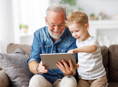 Happy family senior grandfather and boy sitting on couch and watching cartoon on tablet on weekend...