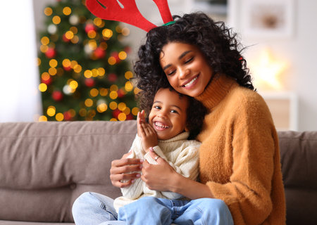 Happy family: cute ethnic boy in sweater playing and embracing with cheerful woman on comfortable couch on Christmas eve at homeの写真素材