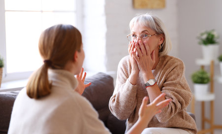 Adult woman gesticulating and sharing amazing news with astonished elderly mother while sitting on sofa at homeの写真素材