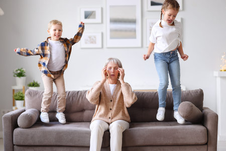 Annoying children jumping on sofa and irritating unrecognizable exhausted woman touching head and...