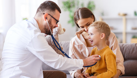 Male medical practitioner showing plush bear and listening lungs of cheerful boy near mother during appointmentの写真素材