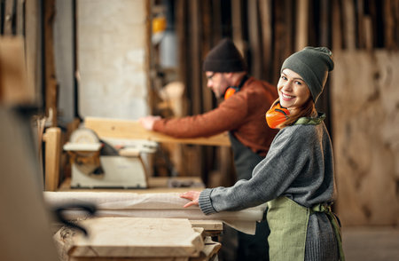 Couple of cheerful carpenters with wooden details in hands working together near workbench in professional workroomの写真素材