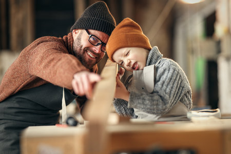 happy family little boy and father in sweaters and hats holding board together while working with wood in modern handicraft studioの写真素材